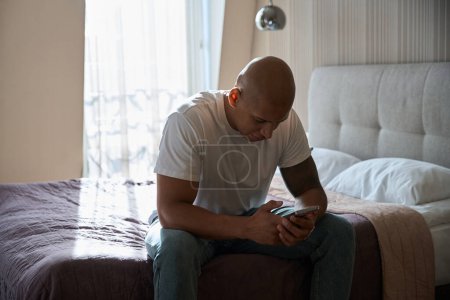 Foto de Hombre afroamericano sentado en la cama y el uso de teléfonos inteligentes en la habitación de hotel en el día soleado. Concepto de descanso, vacaciones y viajes - Imagen libre de derechos