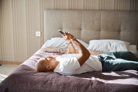 Photo for African american man lying on bed and using smartphone in hotel room at sunny day. Concept of rest, vacation and travelling - Royalty Free Image