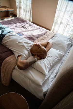Téléchargez les photos : Homme afro-américain les yeux fermés couché et se reposant sur le lit dans la chambre d'hôtel le jour. Concept de repos, vacances et voyages - en image libre de droit