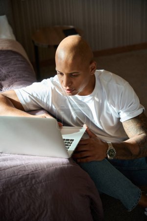 Photo for Focused african american man using laptop on floor by bed in hotel room at daytime. Concept of rest, vacation and travelling - Royalty Free Image