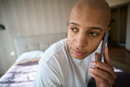 Foto de Primer plano del hombre afroamericano hablando por teléfono móvil en la habitación borrosa del hotel durante el día. Concepto de descanso, vacaciones y viajes - Imagen libre de derechos