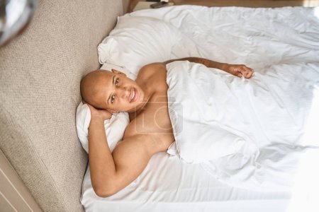Téléchargez les photos : Vue de dessus de l'homme afro-américain couché sur le lit et regardant la caméra dans la chambre d'hôtel à l'heure du matin. Concept de repos, vacances et voyages - en image libre de droit