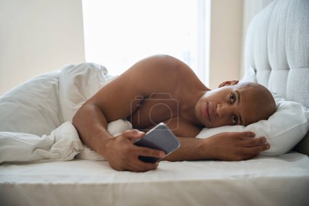 Foto de Joven hombre afroamericano sosteniendo teléfono inteligente y mirando a la cama en la habitación de hotel en la mañana. Concepto de descanso, vacaciones y viajes - Imagen libre de derechos