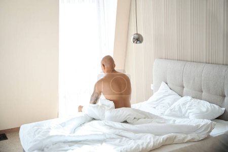 Téléchargez les photos : Jeune homme afro-américain tatoué assis sur le lit dans la chambre d'hôtel à l'heure du matin ensoleillé. Concept de repos, vacances et voyages - en image libre de droit