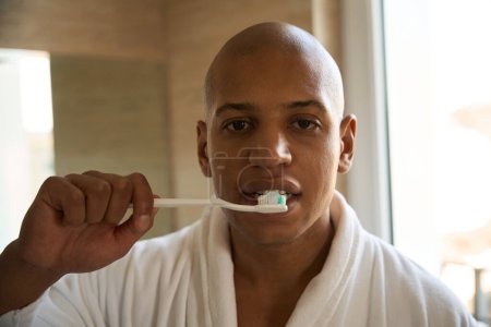 Foto de Retrato de un joven afroamericano cepillándose los dientes con cepillo de dientes en el baño por la mañana. Concepto de procedimientos matutinos e higiene - Imagen libre de derechos