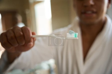 Foto de Enfoque selectivo del cepillo de dientes con pasta de dientes en la mano del hombre afroamericano borroso en el baño por la mañana. Concepto de procedimientos matutinos e higiene - Imagen libre de derechos