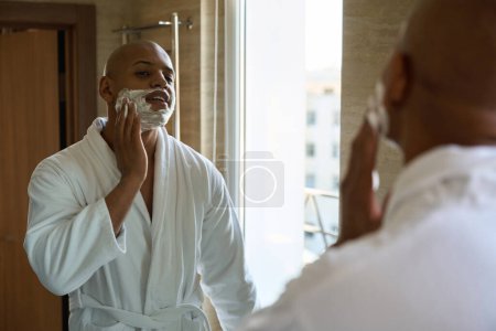 Photo for Young african american man smearing shaving foam on face and looking at himself in mirror in bathroom at morning time. Concept of morning procedures and hygiene - Royalty Free Image