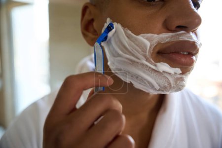 Téléchargez les photos : Visage obscur de jeune homme afro-américain se rasant avec un rasoir dans la salle de bain le matin. Concept de procédures matinales et d'hygiène - en image libre de droit
