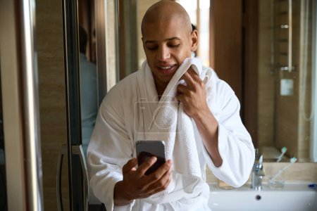 Téléchargez les photos : Jeune homme afro-américain séchage visage avec serviette et regarder smartphone dans la salle de bain à l'heure du matin. Concept de procédures matinales et d'hygiène - en image libre de droit