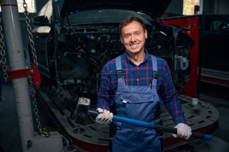 Photo for Portrait of smiling employer in protective costume standing near automobile, holding hammer, ready to work - Royalty Free Image