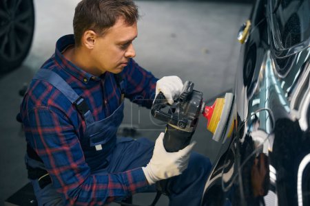 Photo for Mechanic in uniform polishes car detail, assesses damage of hood surface. Surface preparation before painting - Royalty Free Image