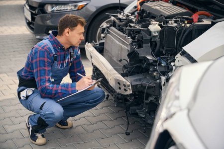 Photo for Young male holding clipboard and performing check of car condition. Standing near automobile. Tire repair - Royalty Free Image