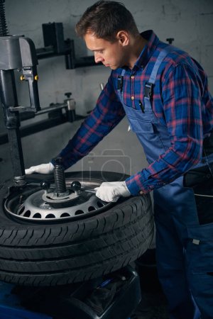 Photo for Mechanic in protective uniform and gloves standing near wheel and checking condition, performing repair - Royalty Free Image