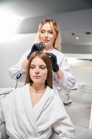 Female european beautician doing trichoscopy with modern trichoscope of young caucasian woman hair in blurred beauty salon. Concept of hair care