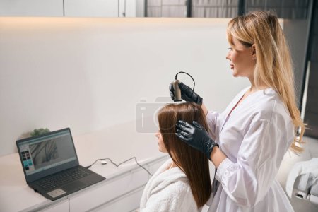 Side view of female cosmetologist doing trichoscopy with modern trichoscope and watching results on laptop of woman hair in blurred beauty salon. Concept of hair care