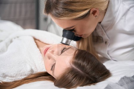 Female beautician examining face skin with dermatoscope of young woman in blurred beauty salon. Concept of face skin care