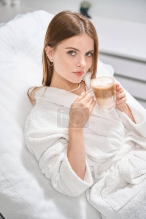 Téléchargez les photos : Jeune femme caucasienne avec une tasse de cappuccino couchée sur le canapé et regardant la caméra dans un salon de beauté flou. Concept de soin de la peau corporelle - en image libre de droit