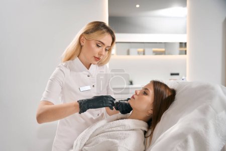 Foto de Vista lateral de una cosmetóloga europea haciendo inyección de belleza facial con jeringa de mujer joven caucásica en un sofá en un salón de belleza borroso. Cuidado, elevación y rejuvenecimiento de la piel facial - Imagen libre de derechos