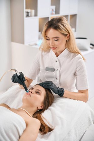 Téléchargez les photos : Femme cosmétologue européenne faisant hydra thérapie faciale de la jeune femme caucasienne sur le canapé dans le salon de beauté floue. Concept de soin, lifting et rajeunissement de la peau du visage - en image libre de droit