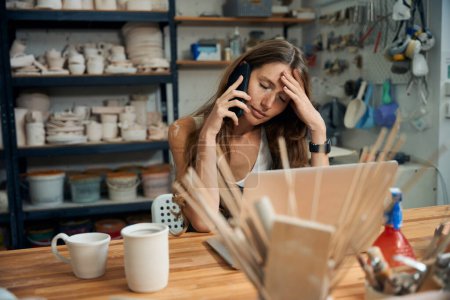 Téléchargez les photos : Fatiguée femme épuisée artiste céramique travaillant sur ordinateur portable et parlant sur téléphone mobile - en image libre de droit
