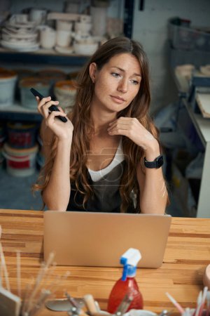Photo for Woman potter holding mobile phone using laptop for working in her creative art studio - Royalty Free Image