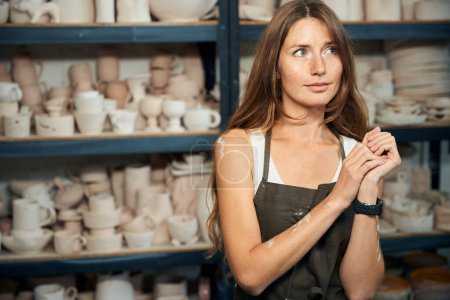 Téléchargez les photos : Pensive femme rêveuse artiste céramiste posant dans son atelier avec des produits en argile sur fond - en image libre de droit