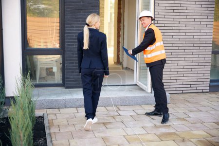 Photo for Back view photo of lady client entering house while male architect wearing helmet and safety vest opening door for her - Royalty Free Image