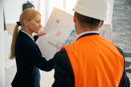 Téléchargez les photos : Femme d'affaires amusée et ingénieur technique debout dans la pièce tout en regardant le plan du bâtiment et en discutant de rénovation - en image libre de droit