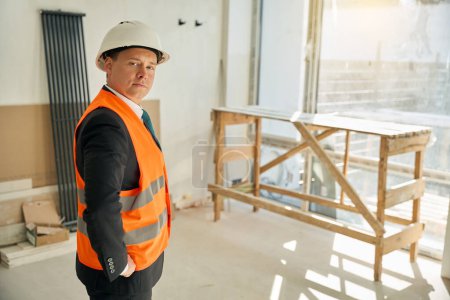 Photo for Waist up picture of male architect wearing helmet and putting hands on hips while standing in room with unfinished renovation - Royalty Free Image