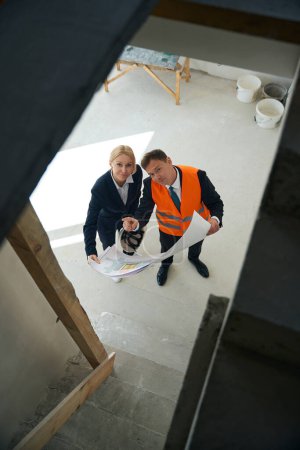 Foto de Ingeniero de construcción masculino y mujer de negocios de pie en la habitación, mientras que el primero apunta a algo al revés y el segundo mirando hacia arriba - Imagen libre de derechos