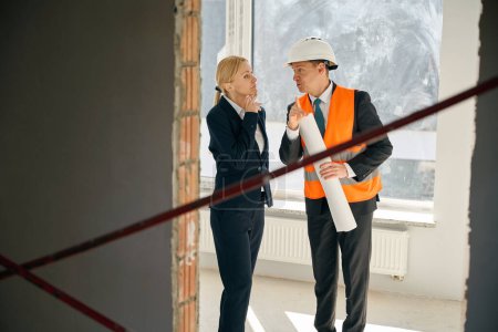 Foto de Gerente de construcción sosteniendo plan enrollado y diciendo algo a los inversores, mientras que los dos de pie en la habitación incompleta - Imagen libre de derechos