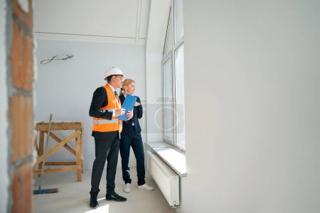 Foto de Ingeniero de construcción sonriente sosteniendo el portapapeles en la mano mientras está de pie con la empleadora cerca de la ventana en la habitación sin terminar - Imagen libre de derechos