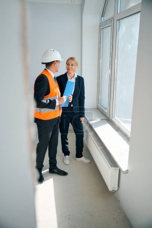 Foto de Foto de larga duración de la alegre mujer inversora e ingeniera de construcción masculina teniendo una charla mientras está de pie en la habitación cerca de la ventana - Imagen libre de derechos