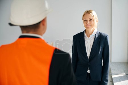 Foto de Foto de la cintura hacia arriba de la mujer de negocios feliz en traje de pie delante del hombre con casco y chaleco de seguridad - Imagen libre de derechos