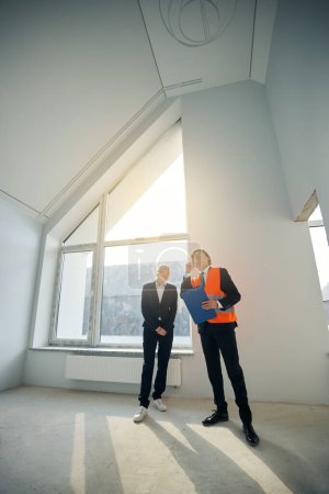 Téléchargez les photos : Homme entrepreneur pointant du doigt le plafond tout en se tenant dans une pièce vide près du client féminin qui regarde vers le haut - en image libre de droit