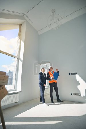 Foto de Hombre capataz señalando el dedo hacia arriba mientras que la mujer cliente mirando el techo cuando ambos de pie en la habitación vacía - Imagen libre de derechos