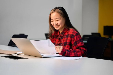 Photo for Young smiling asian female IT employee reading business paper at table in coworking office. Concept of modern freelance or remote work - Royalty Free Image