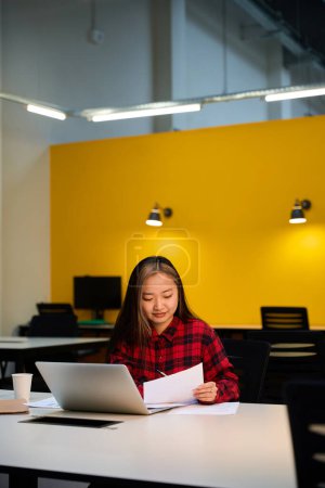 Téléchargez les photos : Jeune asiatique concentrée employée informatique écrivant sur papier tout en utilisant un ordinateur portable au bureau dans le bureau de coworking. Concept de travail indépendant ou à distance moderne - en image libre de droit