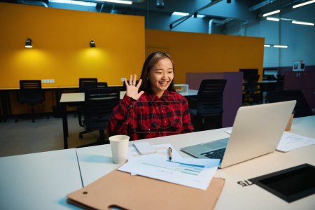 Photo for Young smiling asian female IT employee waving hand during video call on laptop at desk in coworking office. Concept of modern freelance or remote work - Royalty Free Image