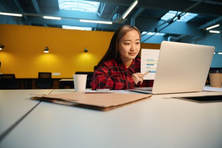 Photo for Young concentrated asian female IT manager showing business graph during video talk on laptop at desk in coworking office. Concept of modern freelance or remote work - Royalty Free Image