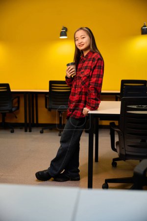 Photo for Side view of young smiling asian female IT employee with coffee cup standing by desk and looking at camera in coworking office. Concept of modern freelance or remote work - Royalty Free Image