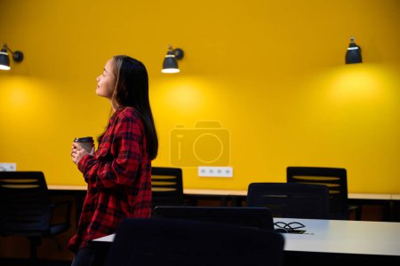 Foto de Vista lateral de la joven y reflexiva mujer asiática empleado de TI con taza de café de pie junto al escritorio y mirando hacia otro lado en la oficina de coworking. Concepto de trabajo independiente o remoto moderno - Imagen libre de derechos
