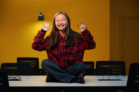 Photo for Front view of young angry asian female IT employee with dog paw model gesticulating sitting on desk in coworking office. Concept of modern freelance or remote work - Royalty Free Image