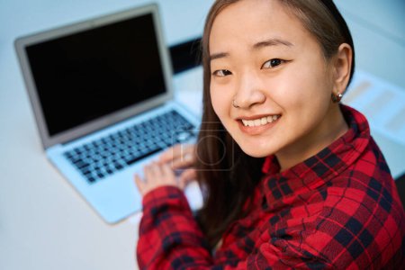Téléchargez les photos : Concentrez-vous sur l'avant-plan de la jeune employée asiatique souriante en informatique qui regarde la caméra tout en travaillant sur un ordinateur portable flou au bureau dans un bureau de coworking. Concept de travail indépendant ou à distance moderne - en image libre de droit