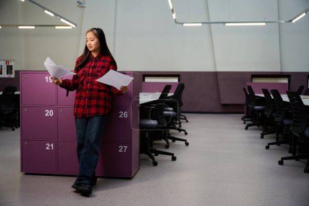 Téléchargez les photos : Jeune asiatique concentrée employée des TI regardant les documents par placard dans le bureau de coworking. Concept de travail indépendant ou à distance moderne - en image libre de droit