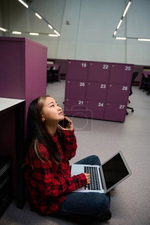Foto de Jóvenes centrado asiático empleado de TI hablando en el teléfono inteligente y el uso de ordenador portátil en el suelo por armario en la oficina de coworking. Concepto de trabajo independiente o remoto moderno - Imagen libre de derechos