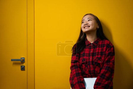 Téléchargez les photos : Jeune asiatique souriante employée de l'informatique avec ordinateur portable regardant loin debout par mur dans le bureau de coworking. Concept de travail indépendant ou à distance moderne - en image libre de droit