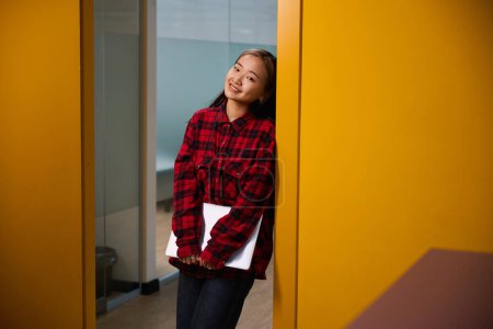 Téléchargez les photos : Jeune asiatique souriante employée de l'informatique avec ordinateur portable regardant la caméra debout dans l'entrée dans le bureau de coworking. Concept de travail indépendant ou à distance moderne - en image libre de droit