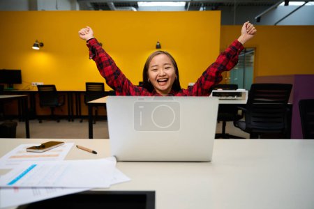 Téléchargez les photos : Jeune asiatique excitée employée des TI avec les mains dans l'air célébrant le succès et regarder ordinateur portable au bureau dans le bureau de coworking. Concept de travail indépendant ou à distance moderne - en image libre de droit