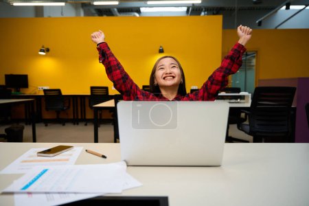 Foto de Joven feliz asiática empleada de TI con las manos en el aire celebrando el éxito en el escritorio con el ordenador portátil abierto en la oficina de coworking. Concepto de trabajo independiente o remoto moderno - Imagen libre de derechos